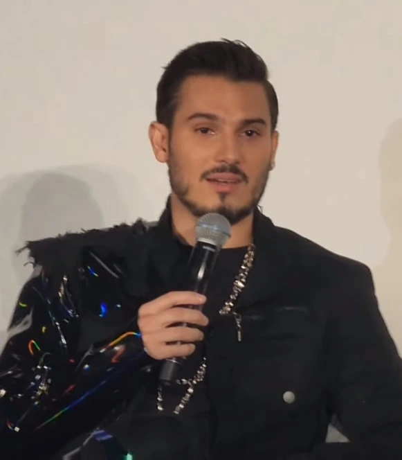 Pipe Bueno holding a microphone during a Q&A appearance, wearing a black outfit and chain necklace, against a plain ligh