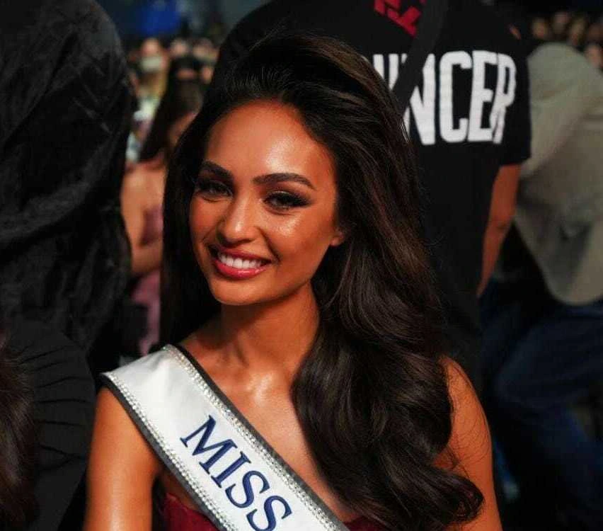 R’Bonney Gabriel smiling in a candid event photo wearing a Miss Universe sash and denim jacket